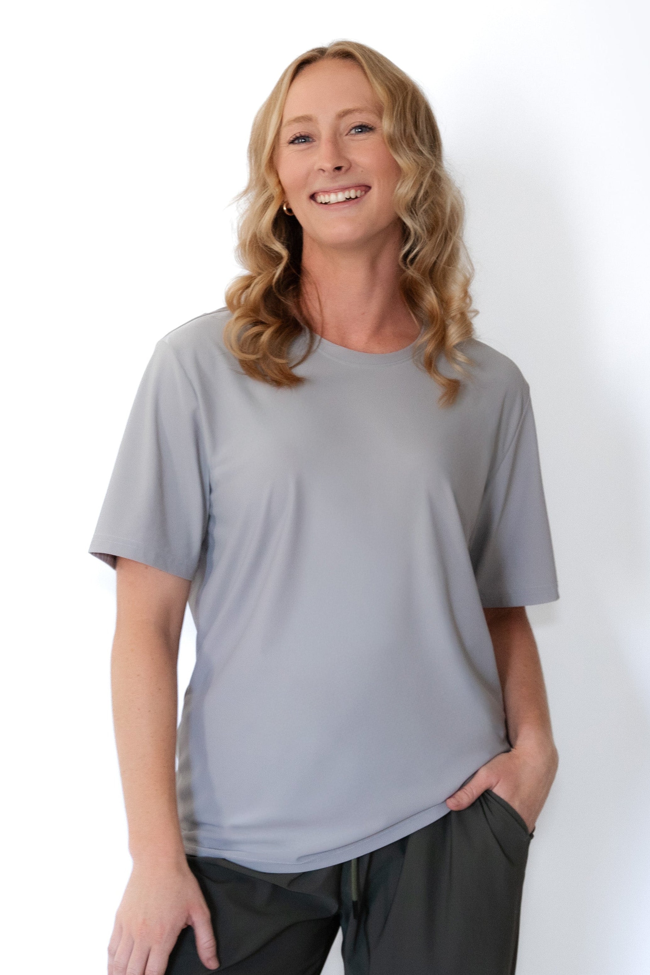 A woman with blonde hair wearing a light gray short-sleeve crewneck t-shirt and dark green pants, standing against a plain white background.