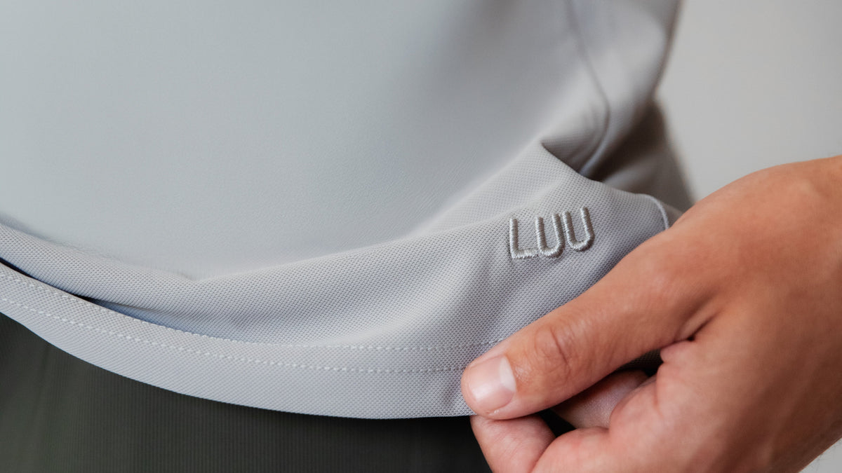 Close-up of a person wearing a light gray athletic top,  pulling the hem of the shirt slightly to reveal a small embroidered logo that reads 'LUU'.